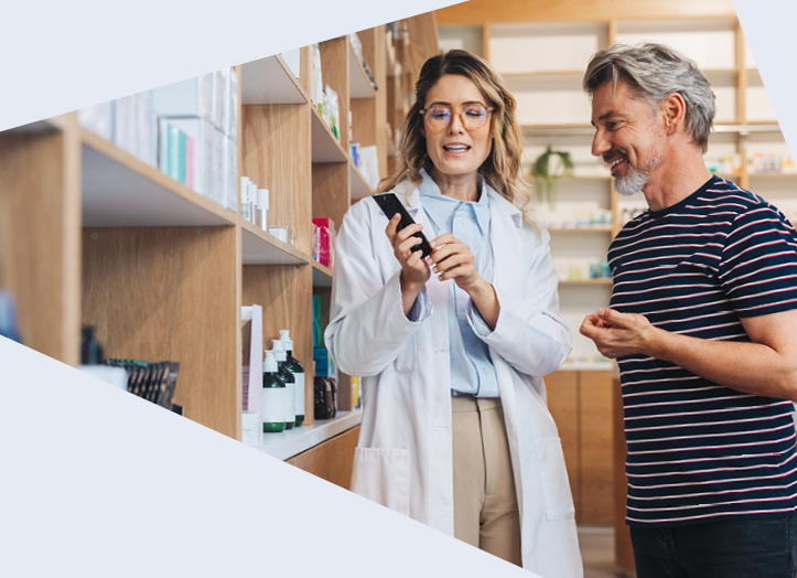 Zwei Personen stehen in einer Apotheke vor einem Regal, eine Person im weißen Kittel hält eine Smartphone und erklärt etwas.