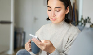 Eine junge Frau sitzt auf einem Sofa mit einer Tasse in der einen Hand und einem Smartphone in der anderen Hand. Sie schaut auf das Smartphone.