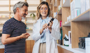 Zwei Personen stehen in einer Apotheke vor einem Regal, eine Person im weißen Kittel hält eine Smartphone und erklärt etwas.