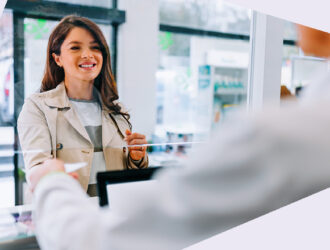 Eine Person steht in einer Apotheke und übergibt ein Rezept an einen Apothekenmitarbeitenden. Dieser steht hinter dem HV und streckt die Hand aus um das Rezept entgegen zu nehmen.