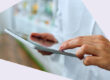 Eine Person in einem weißen Arzt- oder Apothekenkittel hält ein Tablet und tippt darauf. Im Hintergrund sind unscharf Regale mit Produkten zu sehen.