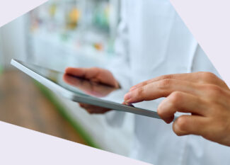 Eine Person in einem weißen Arzt- oder Apothekenkittel hält ein Tablet und tippt darauf. Im Hintergrund sind unscharf Regale mit Produkten zu sehen.