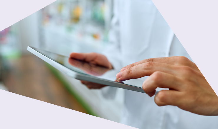 Eine Person in einem weißen Arzt- oder Apothekenkittel hält ein Tablet und tippt darauf. Im Hintergrund sind unscharf Regale mit Produkten zu sehen.