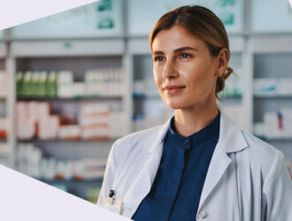 Eine Person in weißem Kittel schaut geradeaus in neutraler Körperhaltung. Im Hintergrund sind Regale mit Arzneimitteln unscharf zu sehen.