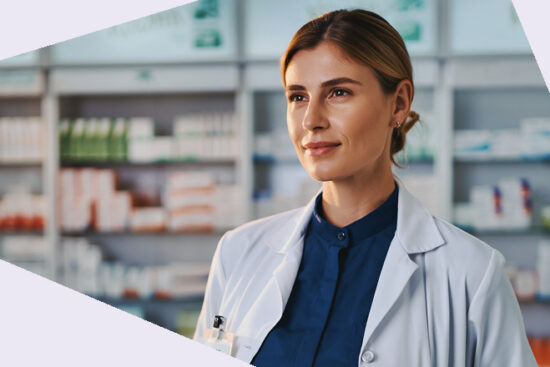 Eine Person in weißem Kittel schaut geradeaus in neutraler Körperhaltung. Im Hintergrund sind Regale mit Arzneimitteln unscharf zu sehen.