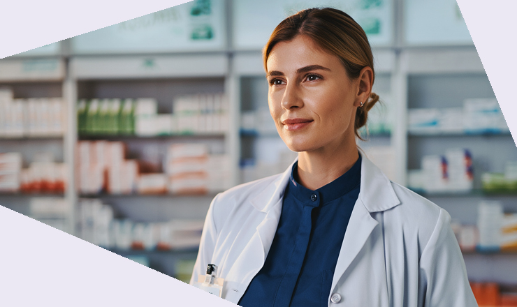 Eine Person in weißem Kittel schaut geradeaus in neutraler Körperhaltung. Im Hintergrund sind Regale mit Arzneimitteln unscharf zu sehen.