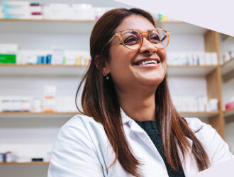 Person mit langen braunen Haaren im weißen Kittel steht vor Regalen voller Arzneimittel.