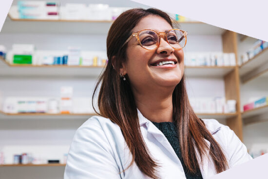 Person mit langen braunen Haaren im weißen Kittel steht vor Regalen voller Arzneimittel.
