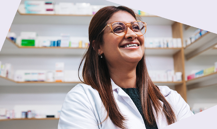 Person mit langen braunen Haaren im weißen Kittel steht vor Regalen voller Arzneimittel.