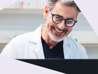 Mann im weißen Kittel steht hinter einem Bildschirm, blickt lachend auf den Bildschirm. Hintergrund zeigt Regale.