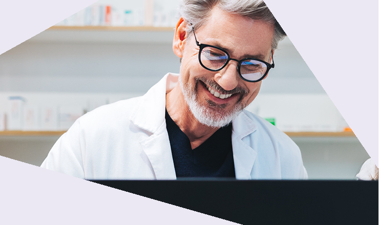 Mann im weißen Kittel steht hinter einem Bildschirm, blickt lachend auf den Bildschirm. Hintergrund zeigt Regale.