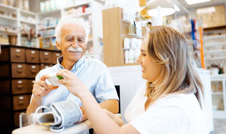 Ein älterer Mann und eine Apothekerin sitzen lächelnd neben einem Apothekentresen. Die Apothekerin zeigt ihm ein Medikament, parallel misst sie Blutdruck bei dem älteren Mann.