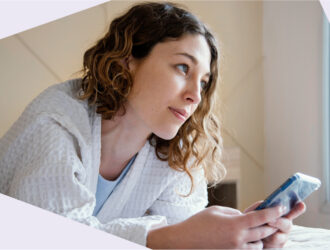 Eine junge Frau im weißen Bademantel liegt auf einem Bett, hält ein Smartphone in der Hand und schaut nachdenklich aus dem Fenster.