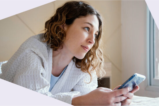 Eine junge Frau im weißen Bademantel liegt auf einem Bett, hält ein Smartphone in der Hand und schaut nachdenklich aus dem Fenster.