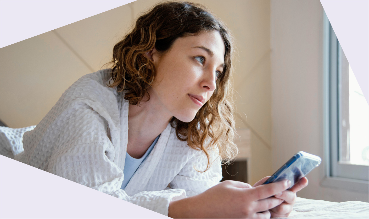 Eine junge Frau im weißen Bademantel liegt auf einem Bett, hält ein Smartphone in der Hand und schaut nachdenklich aus dem Fenster.